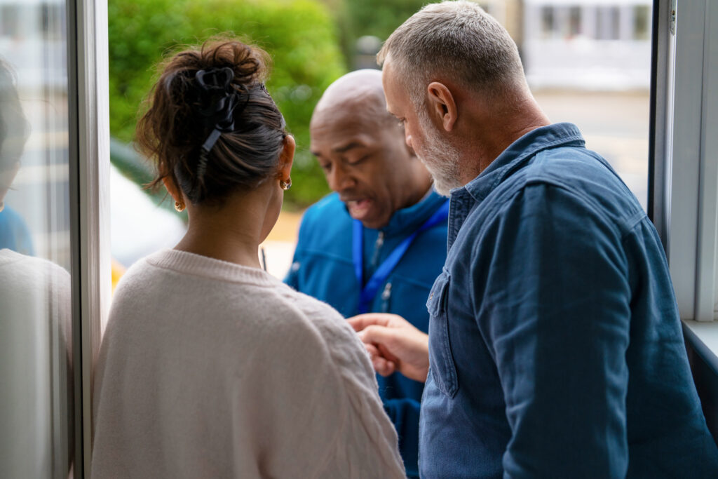 Two individuals are engaged in a conversation with a man standing at the door. A car is visible in the background on a sunny afternoon.
